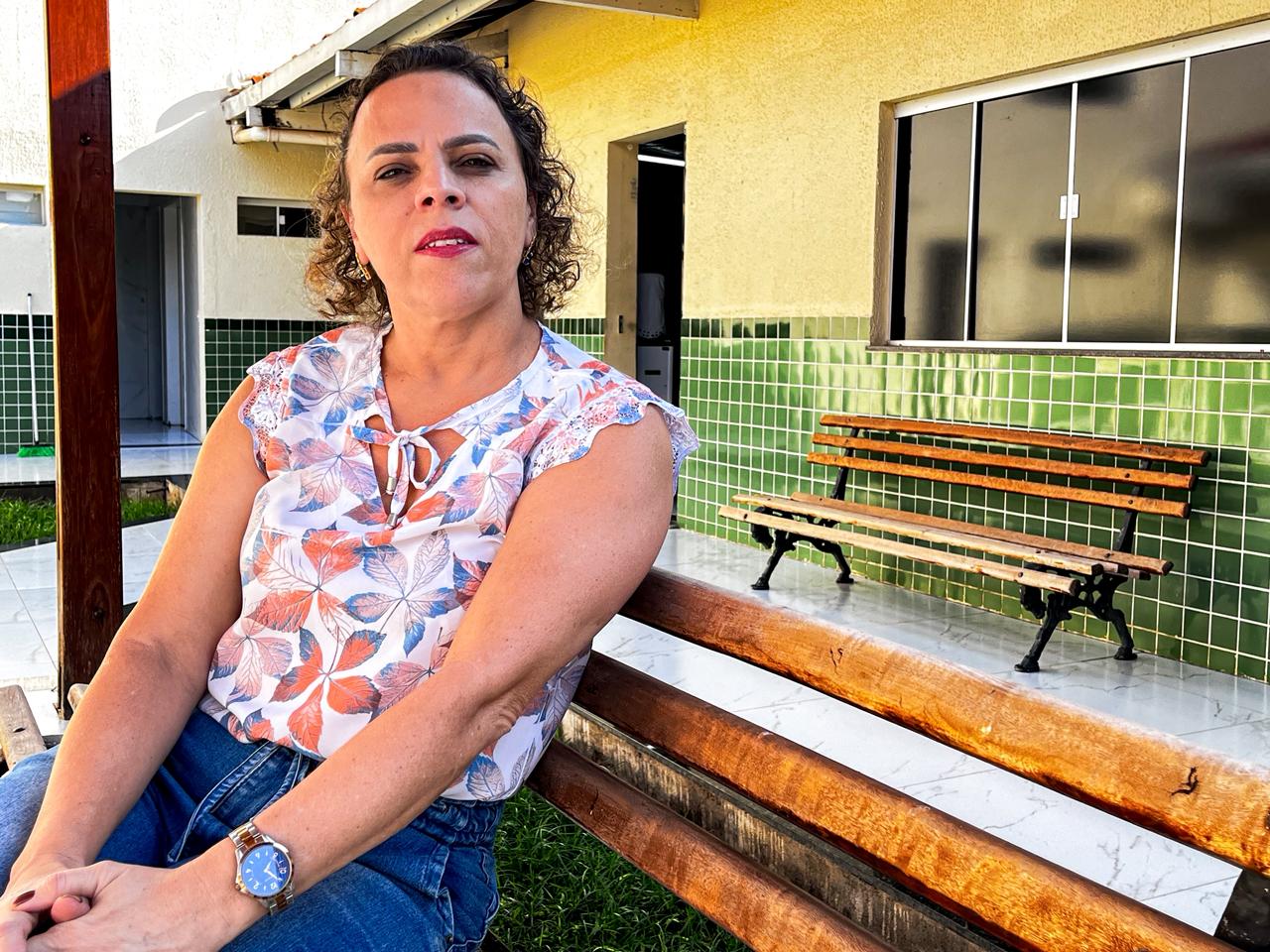 A fotografia, em plano médio, mostra uma mulher branca de cabelos cacheados na altura dos ombros, com mechas mais claras, sentada em um banco de madeira rústica ao ar livre. Ela olha diretamente para a câmera com uma expressão séria e tranquila. Ela veste uma blusa branca sem mangas, estampada com folhas em tons de laranja e azul, e calça jeans azul. Em seu pulso esquerdo, usa um relógio de metal prateado e dourado com mostrador azul escuro. As mãos estão repousadas sobre o colo. Ao fundo, vê-se a fachada de uma construção com a parte inferior da parede revestida por azulejos verdes e a parte superior pintada de amarelo claro. Há uma porta aberta ao centro e uma janela de vidro escuro à direita. Um segundo banco de madeira, vazio, está posicionado próximo à parede, sobre o piso de cerâmica branca. A luz do sol ilumina a cena, vindo da esquerda, criando sombras suaves.