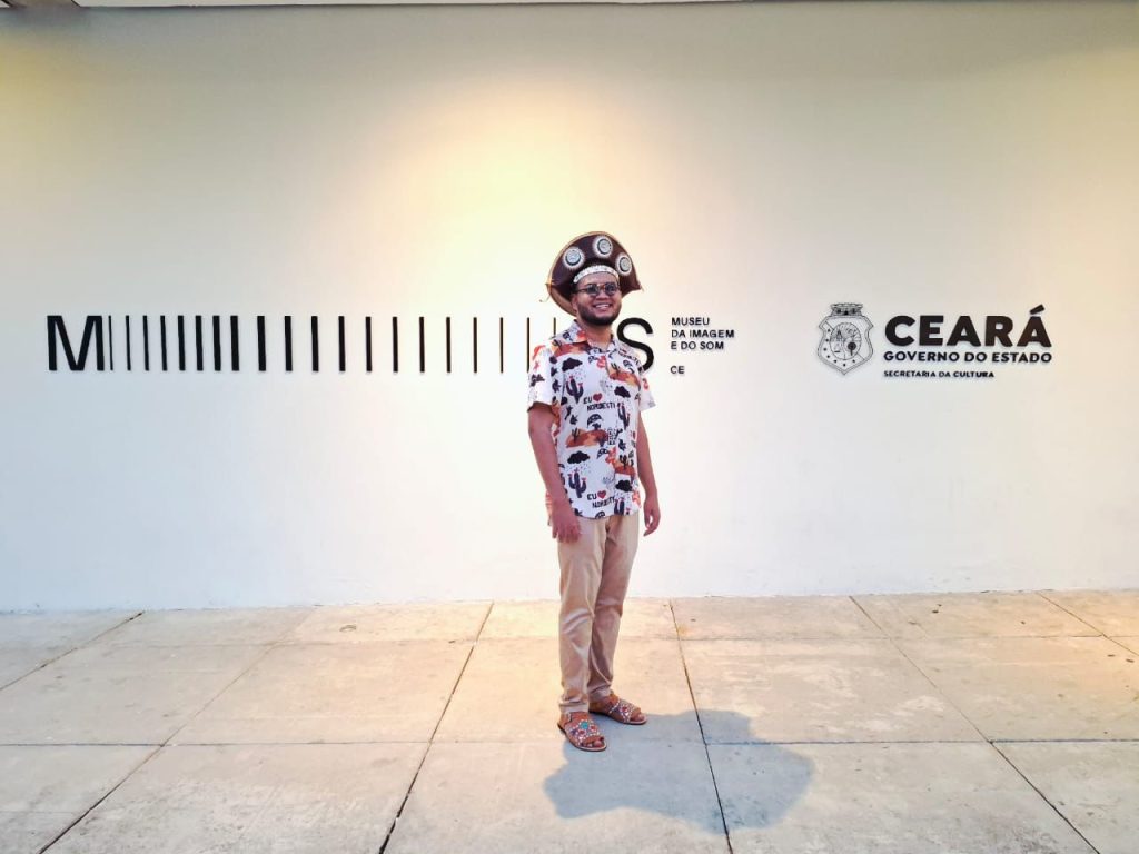 Fotografia de corpo inteiro de Paulo Mateus, o Mateusinho do Acordeon, em pé, no centro da imagem. Ele é um homem de pele parda, barba curta e usa óculos escuros redondos. Ele sorri e veste o mesmo chapéu de couro nordestino da imagem anterior. Sua camisa é branca com estampa colorida de elementos do sertão e frases como "Eu amo o Nordeste", e ele usa calça bege e sandálias de couro marrom com detalhes coloridos. Mateus está em um ambiente interno, bem iluminado, com piso de concreto. Atrás dele, há uma parede branca com inscrições em preto. À esquerda, um código de barras estilizado e o texto "MUSEU DA IMAGEM E DO SOM CE". À direita, o brasão e o texto "CEARÁ GOVERNO DO ESTADO SECRETARIA DA CULTURA".