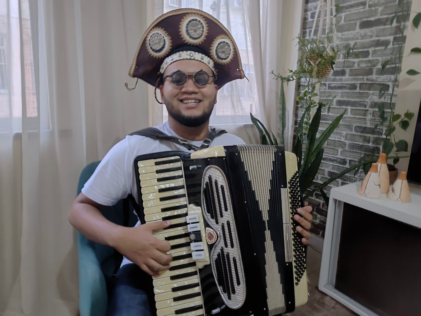Fotografia colorida de Paulo Mateus, o Mateusinho do Acordeon. Ele é um jovem de pele parda, tem barba escura e curta, e sorri abertamente para a câmera. Ele usa óculos com lentes escuras e redondas, e um tradicional chapéu de couro nordestino (estilo cangaceiro) nas cores marrom e bege, decorado com apliques circulares prateados. Mateus veste uma camiseta branca e calça jeans, e está sentado em uma cadeira estofada azul-clara. Ele toca uma sanfona grande, predominantemente preta e creme com uma grade frontal prateada, mantendo as mãos posicionadas sobre as teclas e os botões de baixo do instrumento. O ambiente ao fundo é o interior de uma sala, iluminada pela luz do dia que entra por uma janela com cortinas brancas à esquerda. À direita, há uma parede com papel de parede que imita tijolinhos cinzas, vasos com plantas (incluindo uma espada-de-são-jorge) e um rack branco com três pequenos enfeites rústicos de cerâmica.