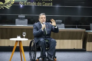 O advogado Emerson Damasceno, presidente da Comissão da OAB-CE, aparece em um auditório oficial. Ele é um homem pardo, de terno cinza e gravata verde, e está sentado em sua cadeira de rodas. Na foto, ele fala ao microfone e gesticula com a mão esquerda. Ao fundo, vê-se uma parede preta com a inscrição dourada "do Estado do Ceará"