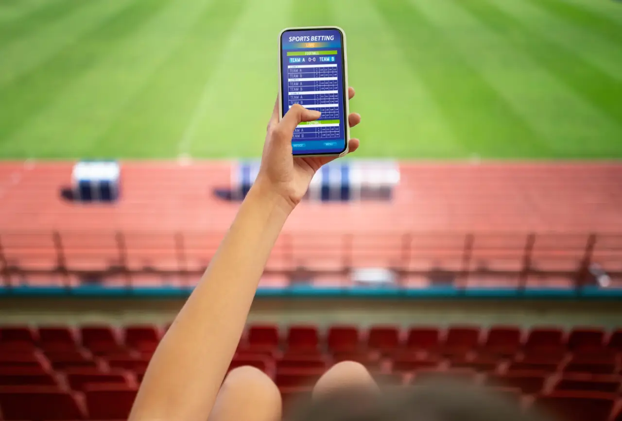 Mão segurando smartphone em arquibancada de estádio mostrando app de bet