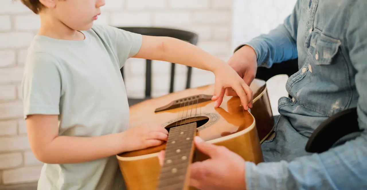 Criança toca em violão segurado por adulto